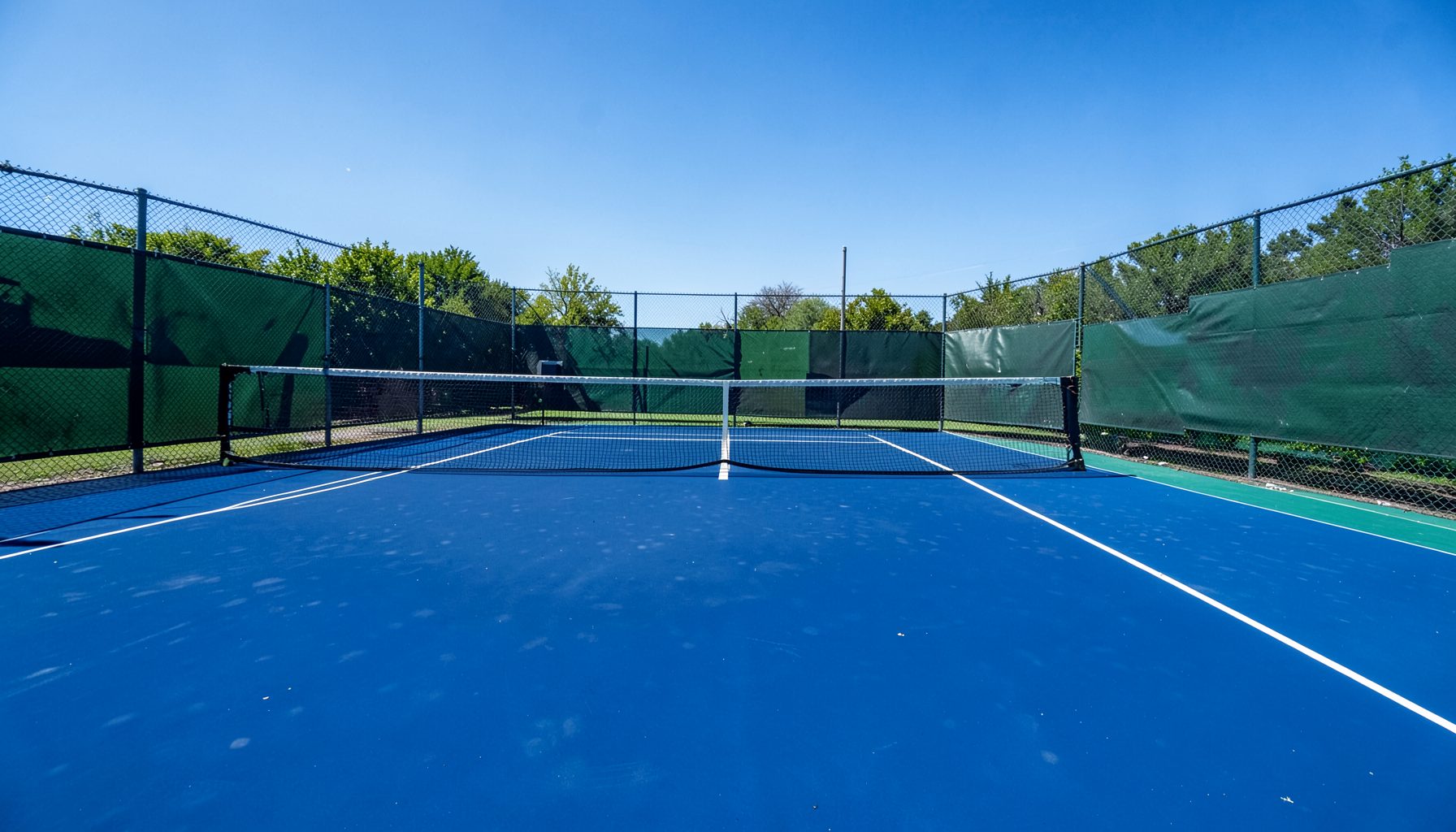 Tennis court with hard court surface stretching toward the net under bright midday sun, deep blue and green tones
