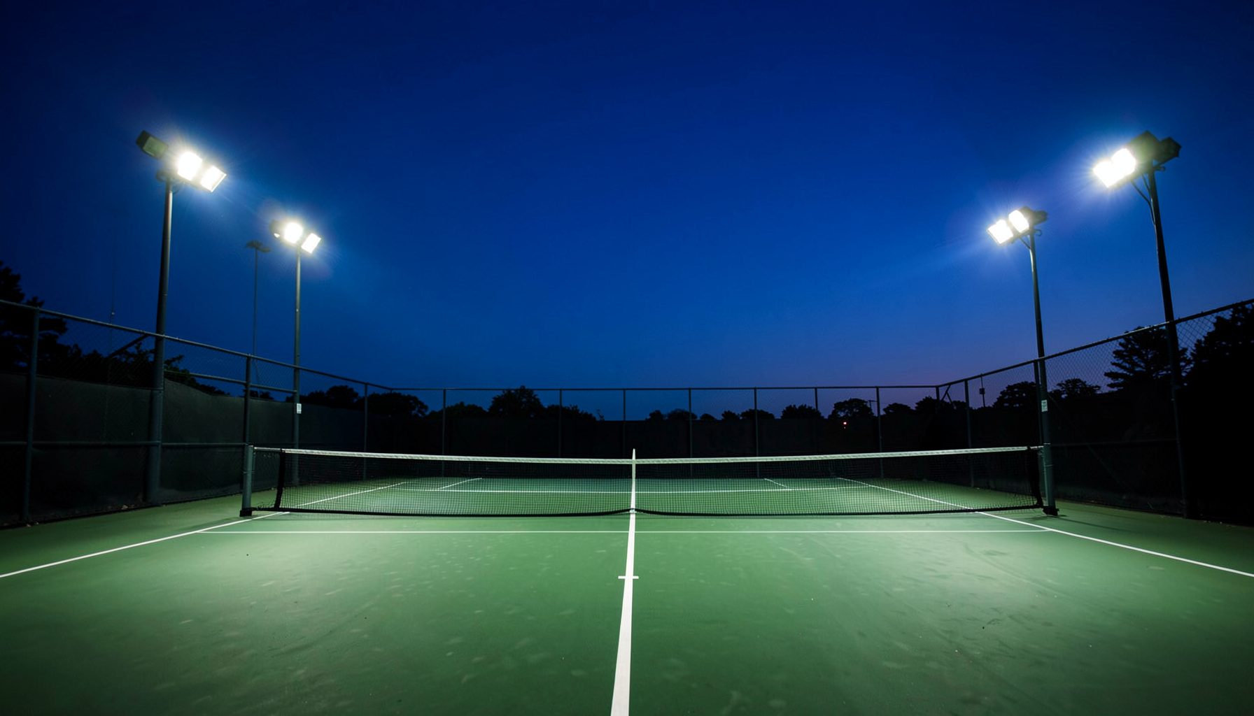 Tennis court at dusk with deep blue sky and bright floodlights casting a dramatic glow across the green court surface